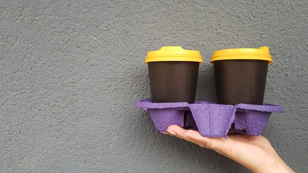 Two glasses of coffee in hand against the background of a blue wall. Takeaway coffee in disposable black cups with yellow lids. Morning coffee outside. Copy space.