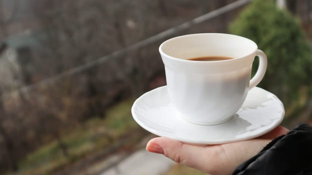 Girl hand holds a cup of white coffee and has a blurred green nature background. Mug in a female hand on a background of mountains