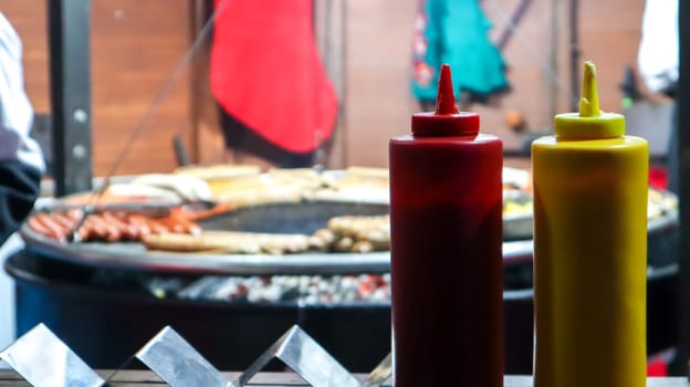 ketchup and mayonnaise in plastic tubes near a street grill in the foreground close-up with selective focus. Barbecue, street food festival. Two cans of meat sauces. Red and yellow container
