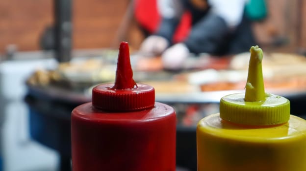 ketchup and mayonnaise in plastic tubes near a street grill in the foreground close-up with selective focus. Barbecue, street food festival. Two cans of meat sauces. Red and yellow container