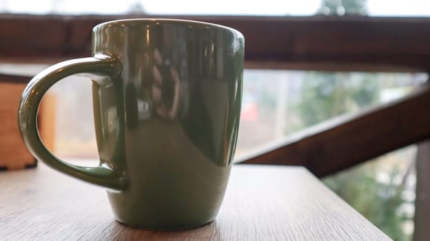 Green cup with tea or coffee on a wooden table on the balcony overlooking the street. Hot drink on the table on the veranda of the summer cafe. Soft focus