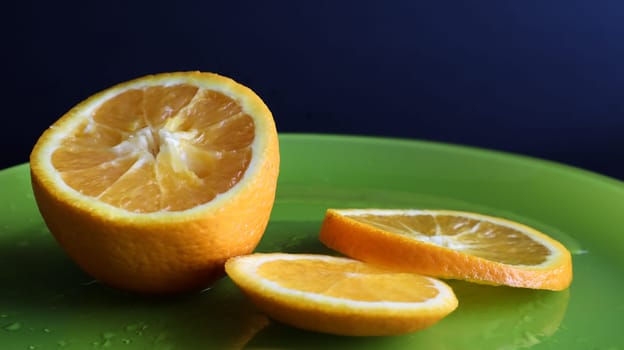 Half an orange and slices on a green glass plate with drops of water, on a table close-up, on a black background. The fruit is multi-nested, multi-seeded, with a thick two-layer peel