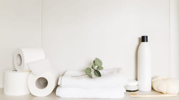 Washbasin in white bathroom, shelf with bath accessories. Hotel cleaning concept. Household concept. Washcloth, shampoo bottle, body cream, white toilet paper rolls, eucalyptus leaf, toothbrush