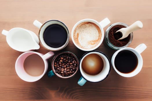Multiple coffee cups and multicolored mugs with americano, espresso, latte and cappuccino, milk jar, roasted coffee beans in glass bottle, wooden spoon and ground coffee in jar on wooden background