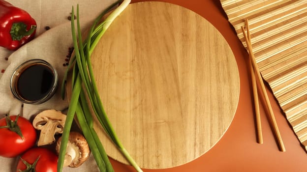 Round wooden board, sauce and various vegetables on the table, wooden stick on a brown background. Flat lay. Copy space in the center of the frame for your text or logo. Recipe and Cooking.