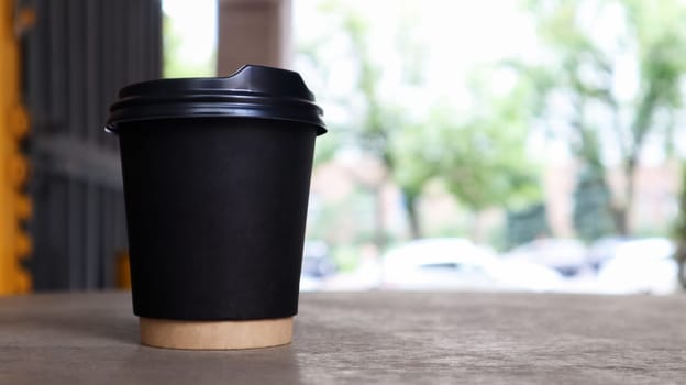 black paper cup without text and logo with coffee with a plastic cover on a wooden table of a street coffee shop in the city. Copy space