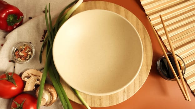 Round wooden board and plate, sauce and various vegetables on the table, wooden stick on a brown background. Flat lay. Copy space in the center of the frame for your text or logo. Recipe and Cooking.