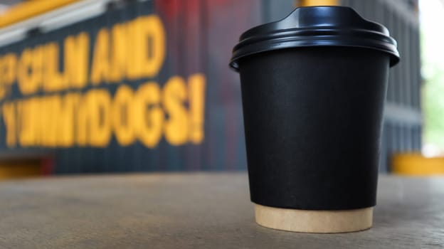 black paper cup without text and logo with coffee with a plastic cover on a wooden table of a street coffee shop in the city. Copy space