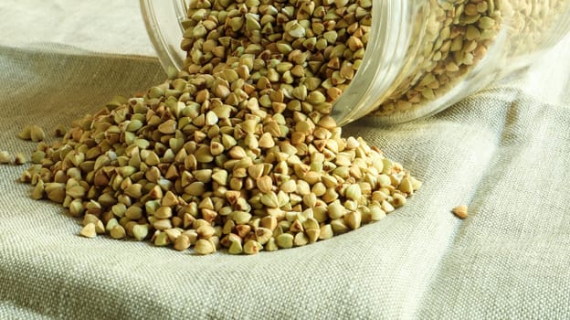 Scattered raw green buckwheat with a glass jar for cereals. Vegan organic food concept. The concept of diet, weight loss, healthy and proper nutrition. Triangle shaped scatter kernels.