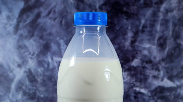 A plastic bottle with a blue cap of fresh regular milk on a dark gray marbled or concrete background. Close-up front view. World milk day concept. Nutrient fluid
