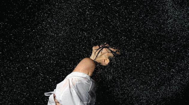 beautiful woman of Caucasian appearance with black hair dances in drops of water on a black background. Woman wearing white shirt