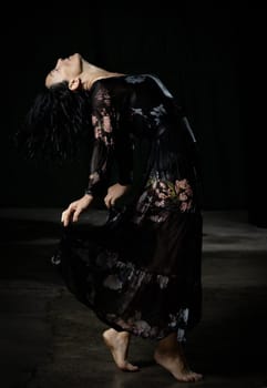 young woman with black hair dances in a black wet long chiffon dress on a dark background. The woman is bent back