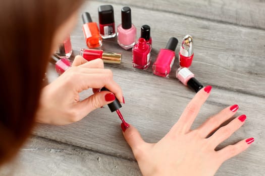 Top view on woman hands, applying red nail polish, with more bottles with varnish in background.