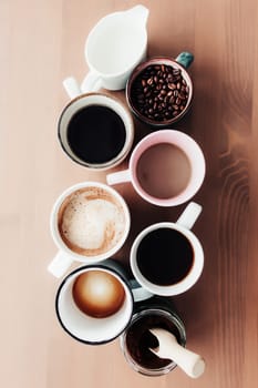 Multiple coffee cups and multicolored mugs with americano, espresso, latte and cappuccino, milk jar, roasted coffee beans in glass bottle, wooden spoon and ground coffee in jar on wooden background