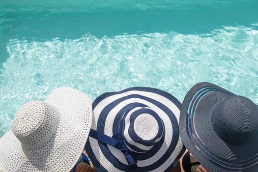 Top view of head of three women wearing straw hat sitting on the edge of the swimming pool. Horizontally.
