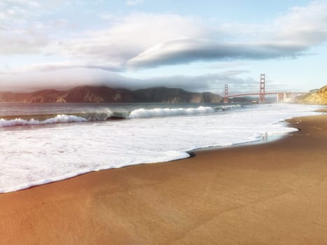 Golden gate bridge view from beach. Cloudy sky in background, waves in foreground.