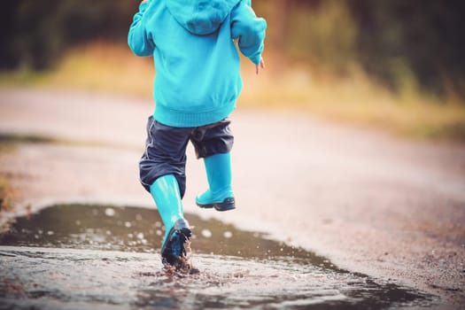 Child walking in wellies and jumping in puddle on rainy weather. Boy under rain in summer outdoors