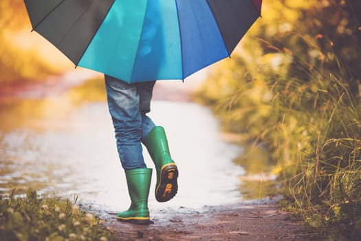 Child walking in wellies in puddle on rainy weather. Boy holding colourful umbrella under rain in summer
