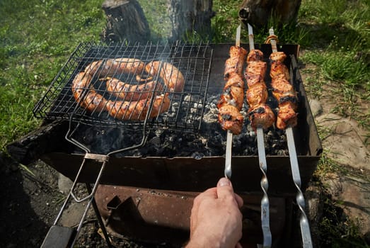 Man preparing shashlik or shish kebab over charcoal. Barbeque meat and sausages or bratwurst on a grill grate in backyard. Grilled meat on metal skewer outdoor. BBQ party or picnic food. Close up shot