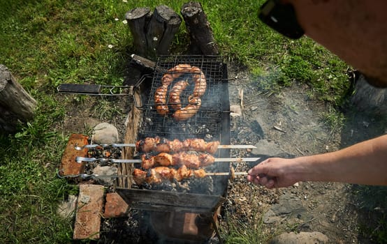 Man preparing shashlik or shish kebab over charcoal. Barbeque meat and sausages or bratwurst on a grill grate in backyard. Grilled meat on metal skewer outdoor. BBQ party or picnic food. Close up shot