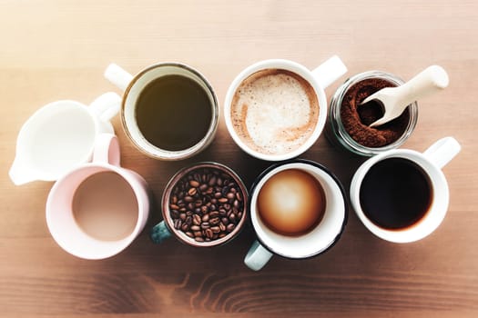 Multiple coffee cups and multicolored mugs with americano, espresso, latte and cappuccino, milk jar, roasted coffee beans in glass bottle, wooden spoon and ground coffee in jar on wooden background