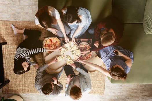 A pizza party, group of friends chatting and happily eating pizza at the table and drinking sweet soda water. Top view from the second floor. Many friends handing out together in green interior house