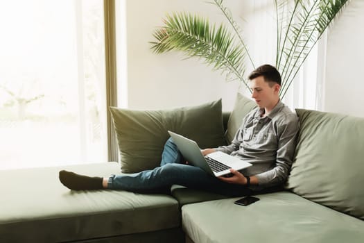 Young guy sitting at green sofa with gray laptop, black phone. Man is holding silver notebook, reading, typing something. Work from home, education concept, watching news and movie. Sunlight in window