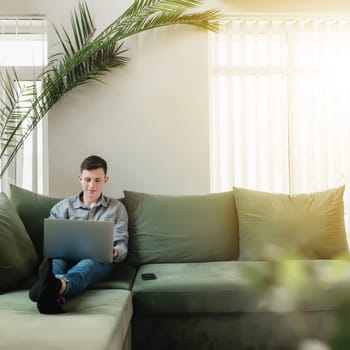 Young guy sitting at green sofa with gray laptop, black phone. Man is holding silver notebook, reading, typing something. Work from home, education concept, watching news and movie. Sunlight in window
