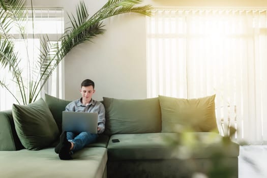 Young guy sitting at green sofa with gray laptop, black phone. Man is holding silver notebook, reading, typing something. Work from home, education concept, watching news and movie. Sunlight in window