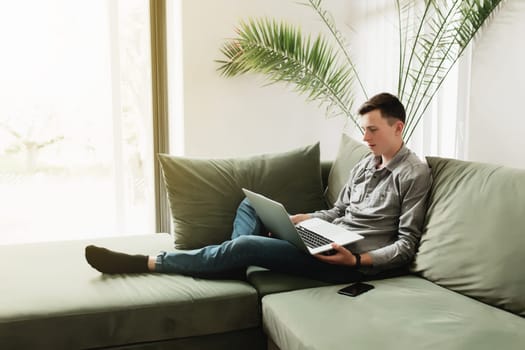 Young guy sitting at green sofa with gray laptop, black phone. Man is holding silver notebook, reading, typing something. Work from home, education concept, watching news and movie. Sunlight in window
