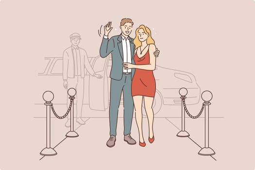 Famous couple waving to camera concept. Young smiling couple stars in elegant clothing standing in hotel entrance waving hands to camera with taxi awaiting behind