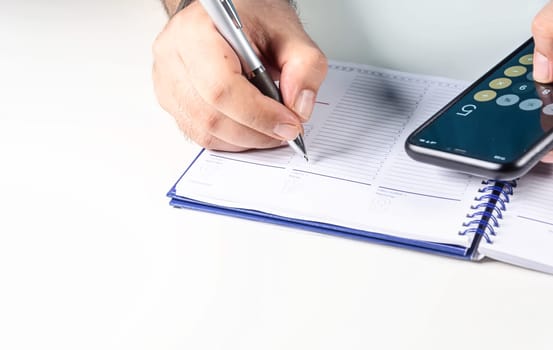 Man doing calculations with his mobile phone calculator and writing in a ring binder notebook with a pen. Budget and economy.