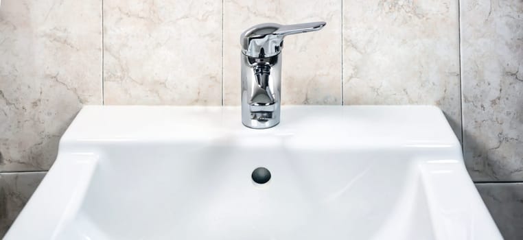 Chromed faucet with lever to mix hot and cold water on a modern white sink in a bathroom with tiled walls. Domestic life and hygiene