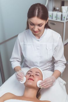 Doctor beautician cleanses face skin with sponge in beauty salon. Perfect cleaning healthcare spa treatment. Skincare beauty spa concept. Model lying on couch with closed eyes in cosmetological clinic