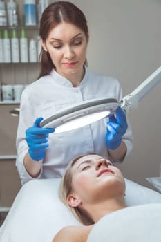 Beautician doctor is examining woman's face with special lamp.