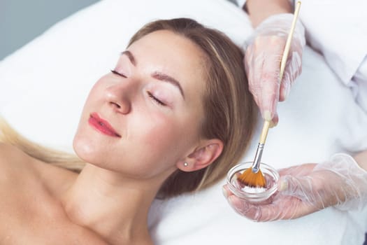 Beautician applying facial mask to the young lady. Blonde caucasian girl at the cosmetology