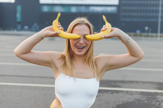 Frivolously and mischievous young woman with braces on teeth holding bananas on her face has fun outdoors. Emotions