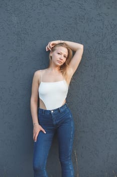 young blonde woman in the white sleeveless t-shirt and blue jeans standing over gray wall background.