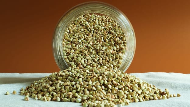 Scattered raw green buckwheat with a glass jar for cereals. Vegan organic food concept. The concept of diet, weight loss, healthy and proper nutrition. Triangle shaped scatter kernels.