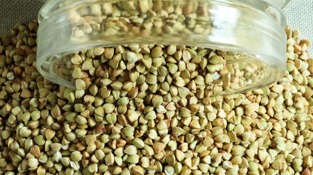 Scattered raw green buckwheat with a glass jar for cereals. Vegan organic food concept. The concept of diet, weight loss, healthy and proper nutrition. Triangle shaped scatter kernels.