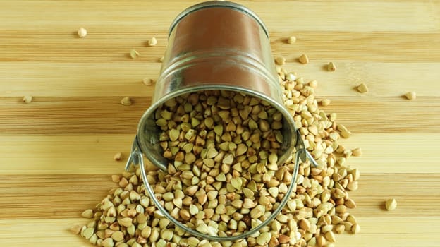 Small metal bucket with raw buckwheat on a board on a brown background. Organic vegetarian food. The concept of a healthy and balanced diet. Dietary nutrition. Copy space. Top view