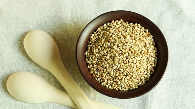 Raw green buckwheat in a brown clay plate and a wooden spoon. Vegan organic food concept. The concept of diet, weight loss, healthy and proper nutrition. Copy space for text. Top view, flat lay.