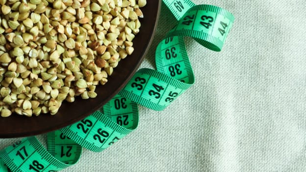 Green measuring tape on a background of raw buckwheat in a dish, concept of diet and healthy nutrition. Vegan organic food concept. Quarantine weight loss. Copy space for text. Top view, flat lay.