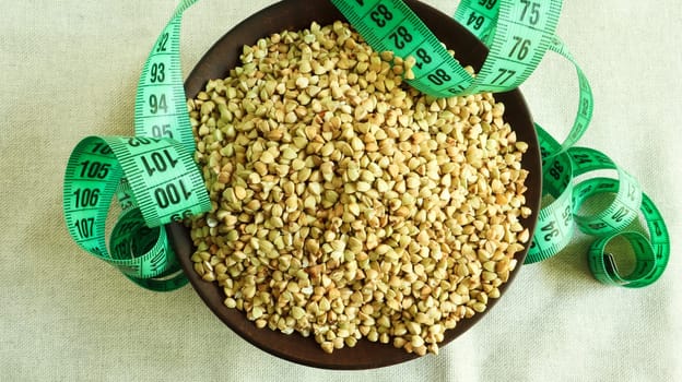 Green measuring tape on a background of raw buckwheat in a dish, concept of diet and healthy nutrition. Vegan organic food concept. Quarantine weight loss. Copy space for text. Top view, flat lay.
