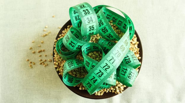 Green measuring tape on a background of raw buckwheat in a dish, concept of diet and healthy nutrition. Vegan organic food concept. Quarantine weight loss. Copy space for text. Top view, flat lay.