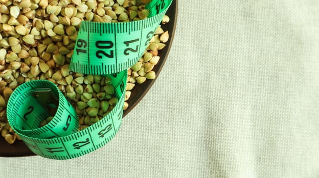 Green measuring tape on a background of raw buckwheat in a dish, concept of diet and healthy nutrition. Vegan organic food concept. Quarantine weight loss. Copy space for text. Top view, flat lay.