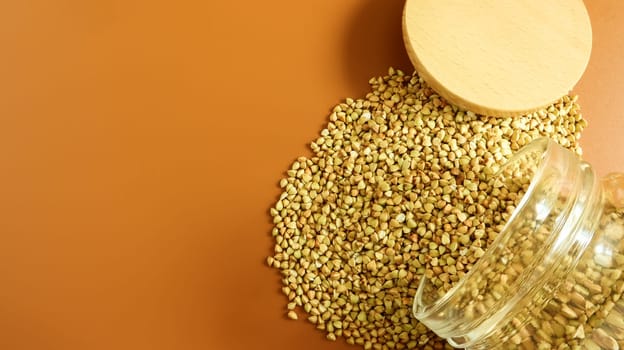 Scattered raw green buckwheat with a glass jar for cereals. Vegan organic food concept. The concept of diet, weight loss, healthy and proper nutrition. Triangle shaped scatter kernels.