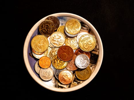 Small money coins in a plate on a black background. Eurocent. Ukrainian hryvnia. Chinese yuan. Currency unit. Small coin. Wealth. Treasure. Background image. Place for text. Poster.