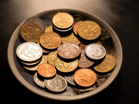 Small money coins in a plate on a black background. Eurocent. Ukrainian hryvnia. Chinese yuan. Currency unit. Small coin. Wealth. Treasure. Background image. Place for text. Poster.