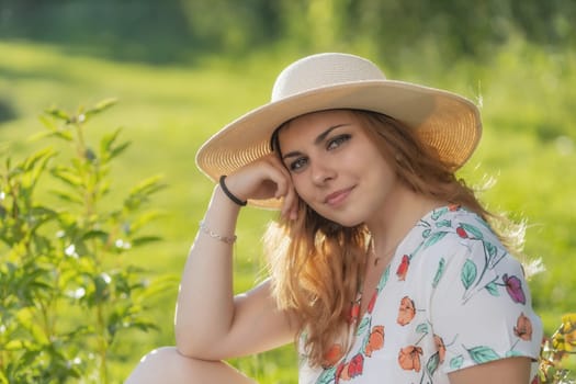Cool young woman wearing straw hat is posing outdoors in summer day. Horizontally.
